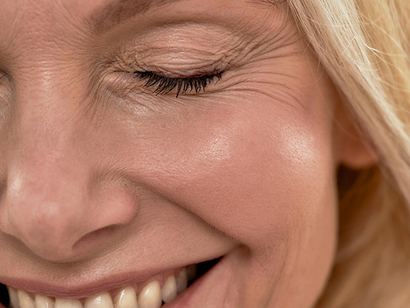 EIne Frau in weißem Bademantel und Stirnband liegt auf einer Liege, zwei Hände in weißen Handschuhen halten ein Wattepad an ihre Wange und Spritzen etwas in die Falten um ihren Mund.